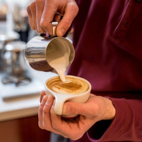 Barista pours cream in a swirly design into small coffee cup