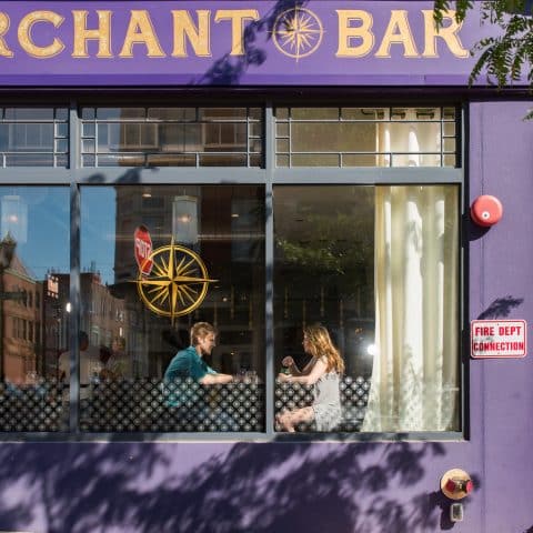 Exterior of purple and yellow Merchant Bar with windows looking in on a man and woman at a table